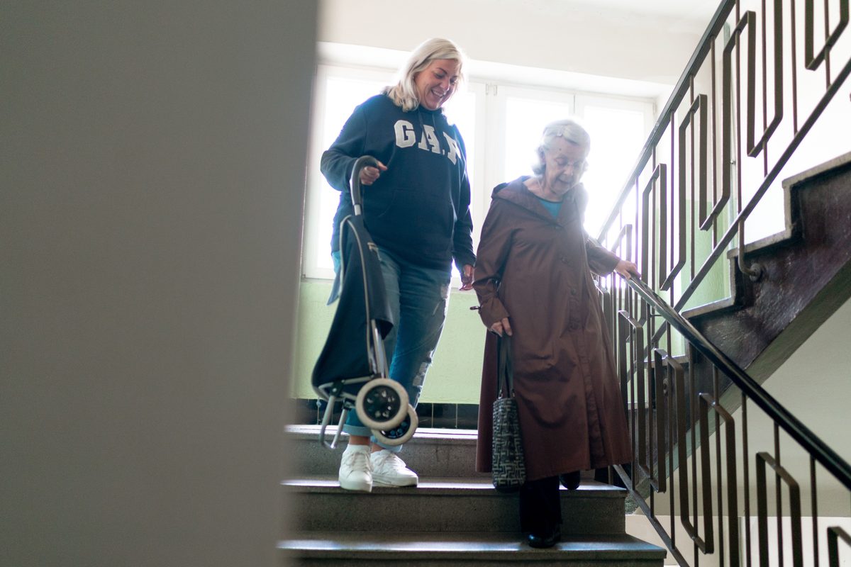 A carer supporting a person walking down a staircase with a walking aid